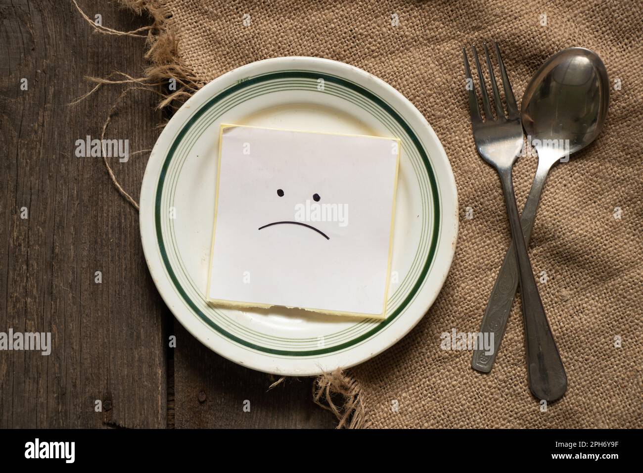 un sorriso triste si trova su un foglio di carta su un piatto accanto a un cucchiaio e una forchetta su uno sfondo marrone burlap, niente da mangiare, crisi Foto Stock