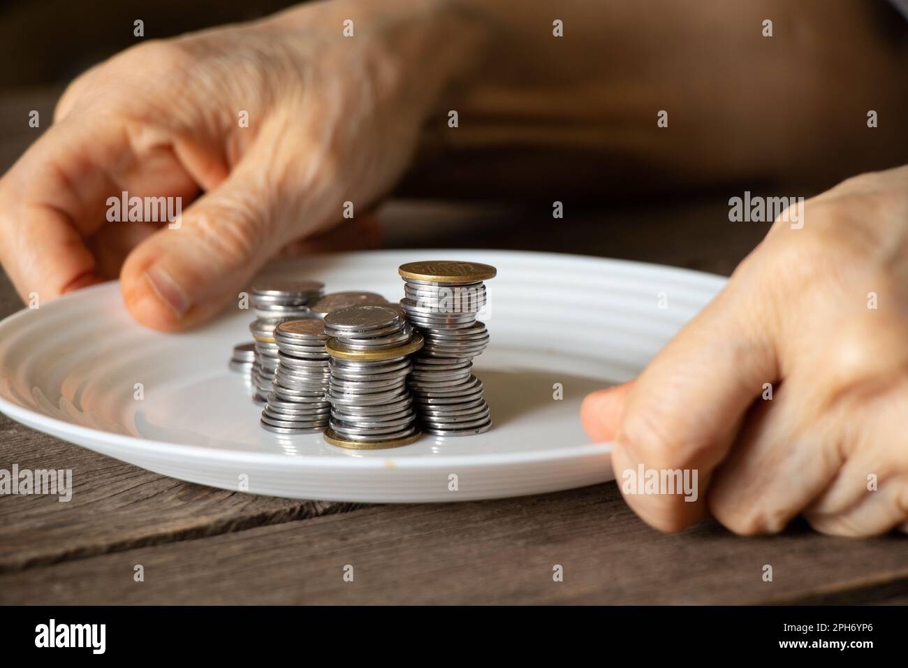 una pila di monete su un piatto bianco su un vecchio tavolo di legno e vecchie mani femminili versarono un piatto a casa in cucina Foto Stock