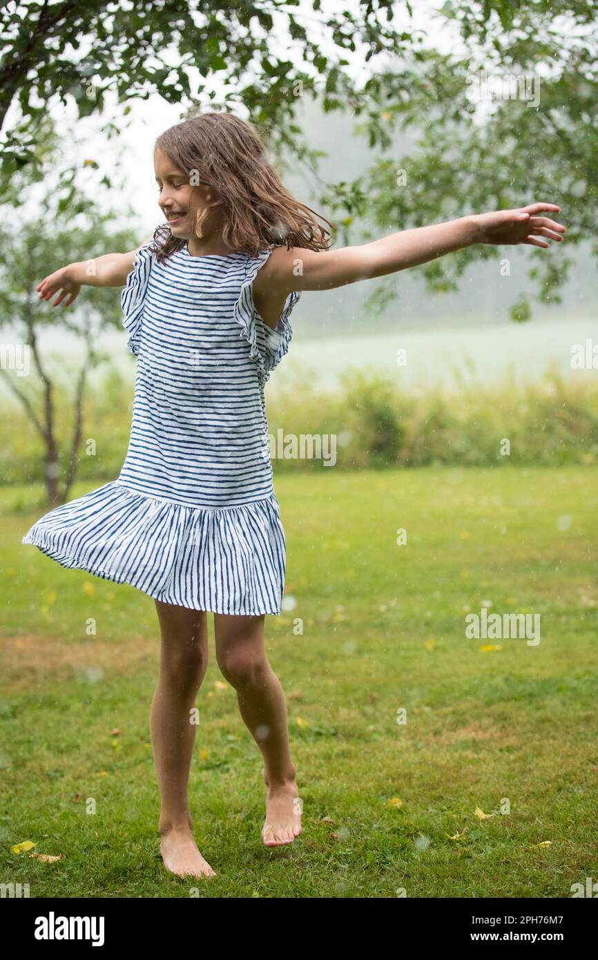 Felice ragazza carina in sundress ballare e correre sul prato verde sotto la pioggia. Bambino a piedi nudi che si gode l'estate all'aperto. Foto Stock