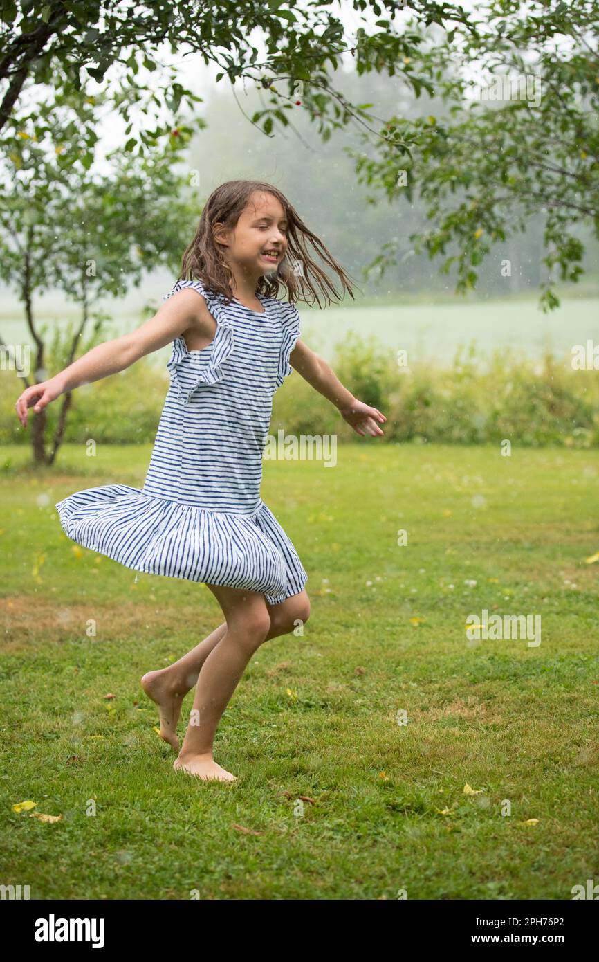 Felice ragazza carina in sundress ballare e correre sul prato verde sotto la pioggia. Bambino a piedi nudi che si gode l'estate all'aperto. Foto Stock