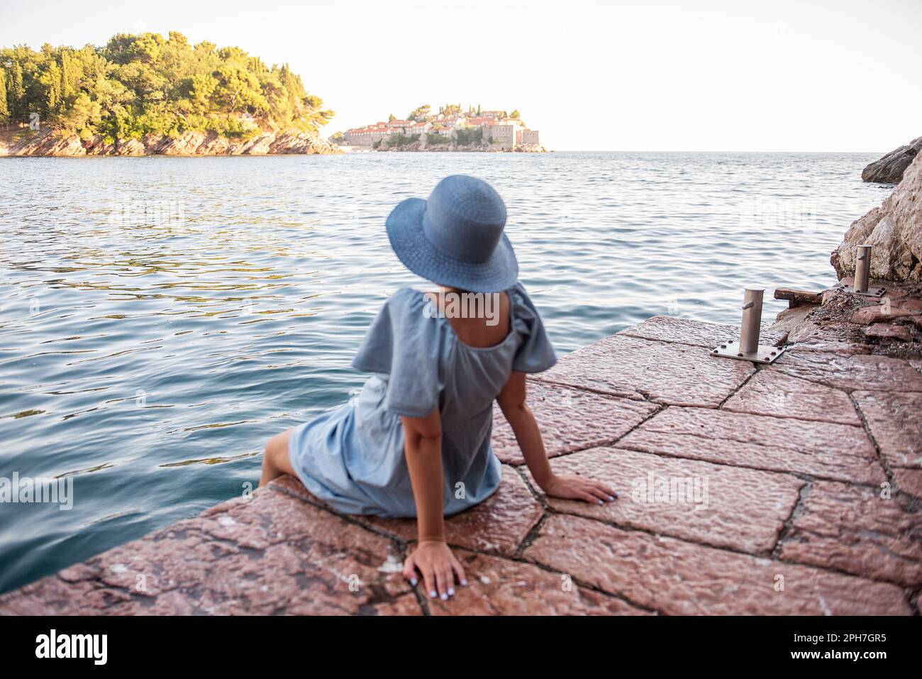 Defocesed donna incinta viaggiatore in denim vestito, cappello di paglia blu siede sul mare, guarda l'isola di Sveti Stefan in Montenegro al tramonto. Touris Foto Stock