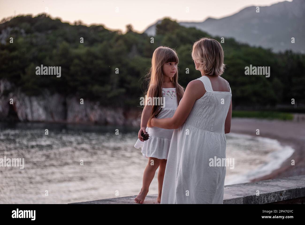 Primo piano ritratto di madre incinta con bambina che cammina mano in mano in abiti bianchi al tramonto sul mare. Assistenza ai bambini. Maternità sana. Foto Stock