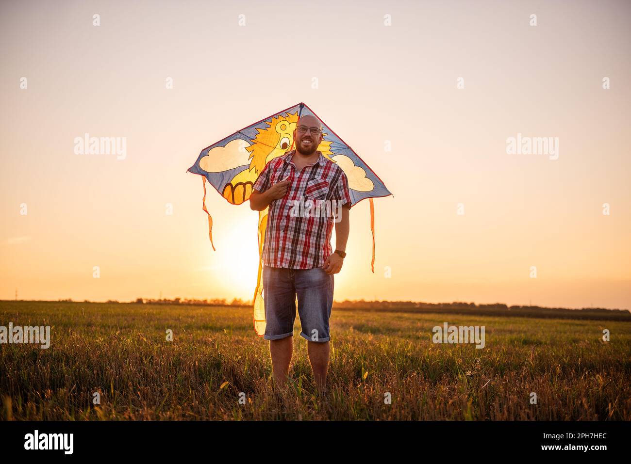 Ai raggi del sole del tramonto, calvo uomo con bicchieri con aquilone nel campo. Il Padre gioca con i bambini nelle aree rurali. Vista attraverso le orecchie del m Foto Stock