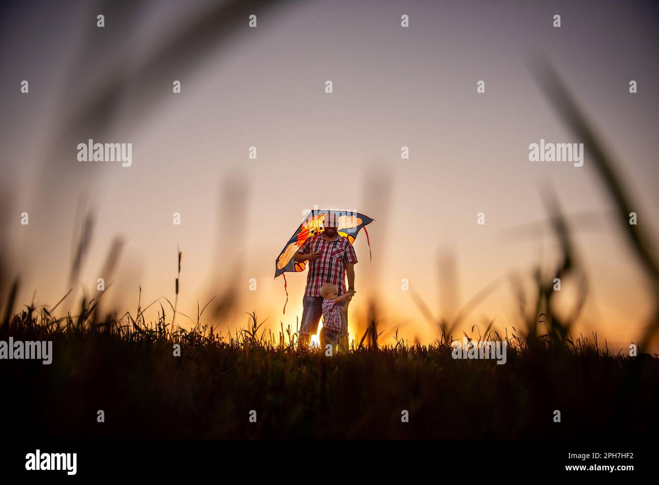 Padre calvo in occhiali in piedi intorno al campo con figlio, tiene dietro aquilone. L'uomo millenario gioca con il ragazzo nelle aree rurali. Viaggi, divertimento di giorno Foto Stock