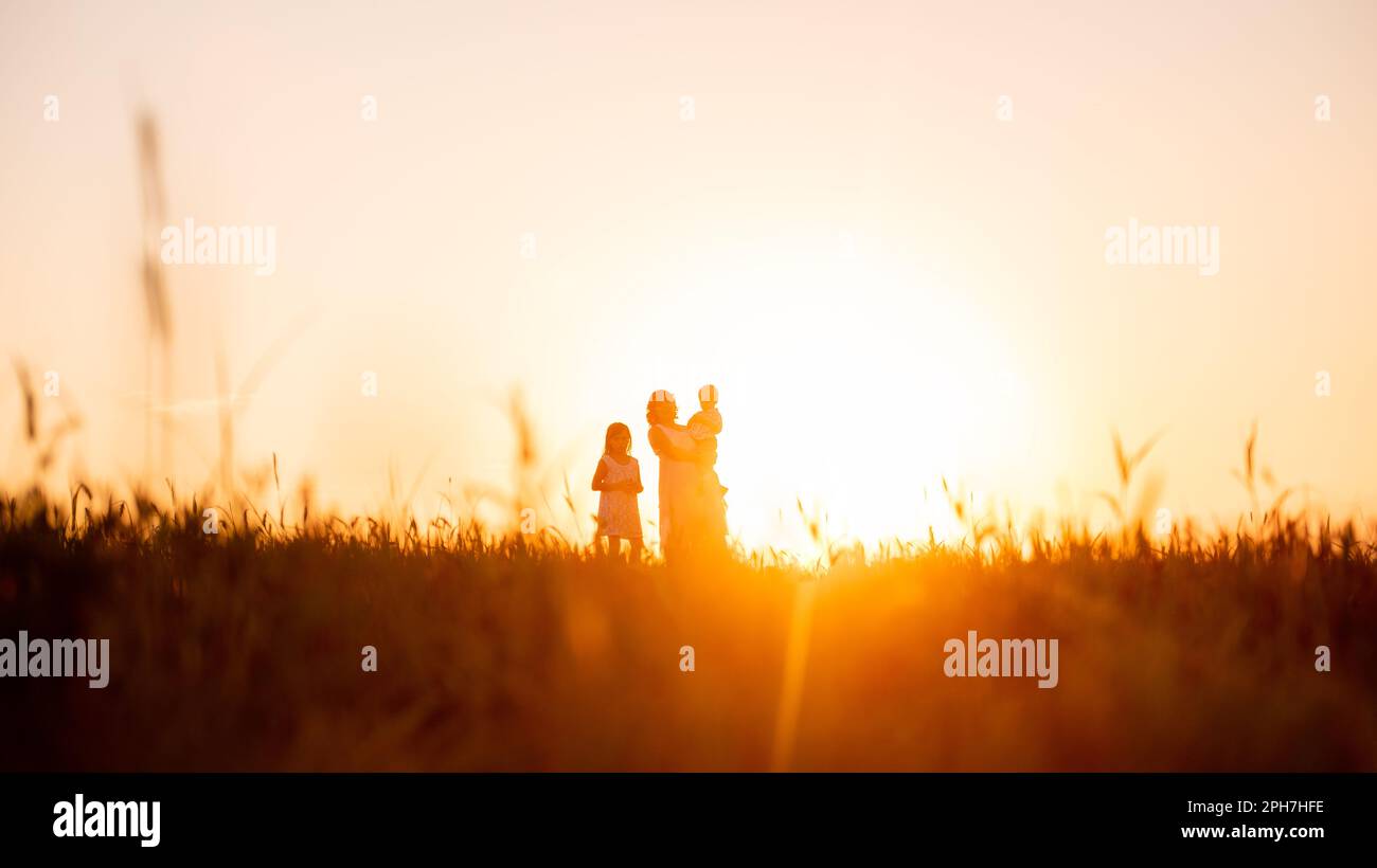 La silhouette delle madri con il figlio in braccio e la figlia al tramonto sul campo. Sfondo naturale arancione. Vista panoramica. Viaggiare con i bambini a. Foto Stock