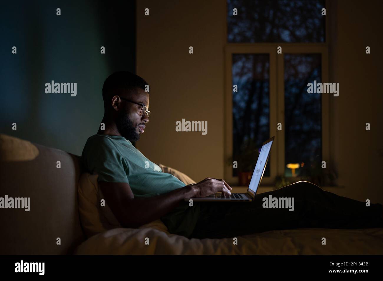 Programmatore africano americano concentrato uomo sdraiato sul letto di lavoro sul laptop di notte. Foto Stock