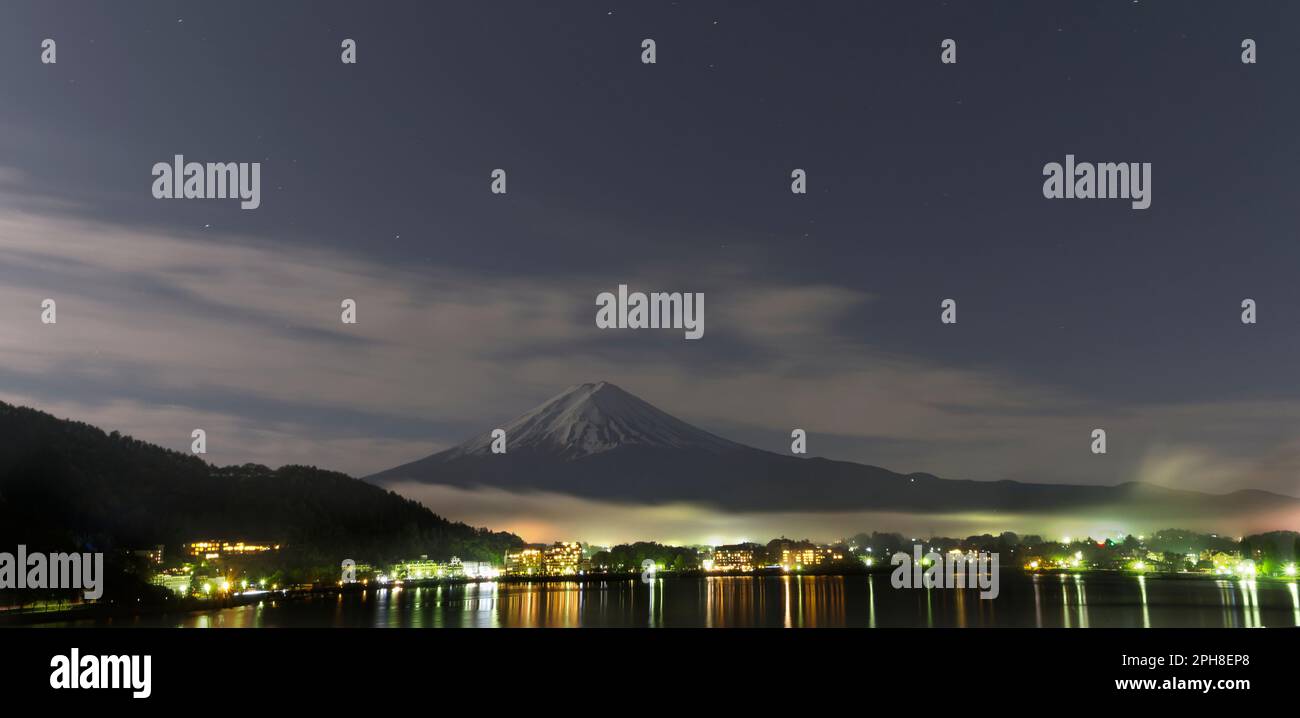 Panorama del lago Kawaguchiko, Monte Fuji. Le luci della città riflettono, specchi sull'acqua calma del lago. Paesaggio notturno con stelle, cielo blu, nuvole, notte ventosa fredda. Foto Stock