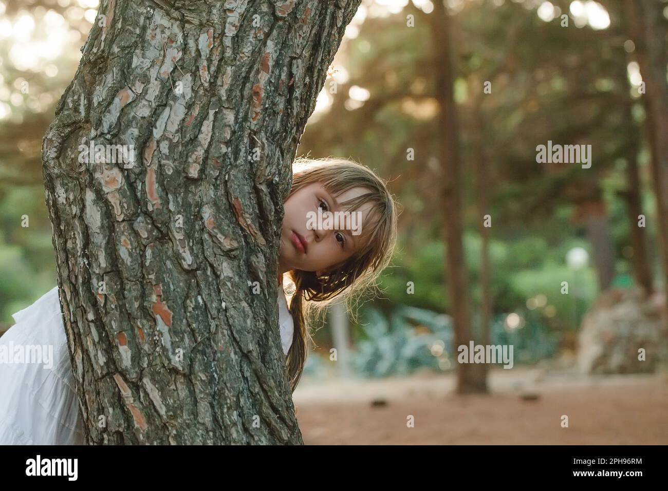 Ritratto di bambina brunetta in abiti bianchi che sbircia da dietro l'albero nel parco Foto Stock
