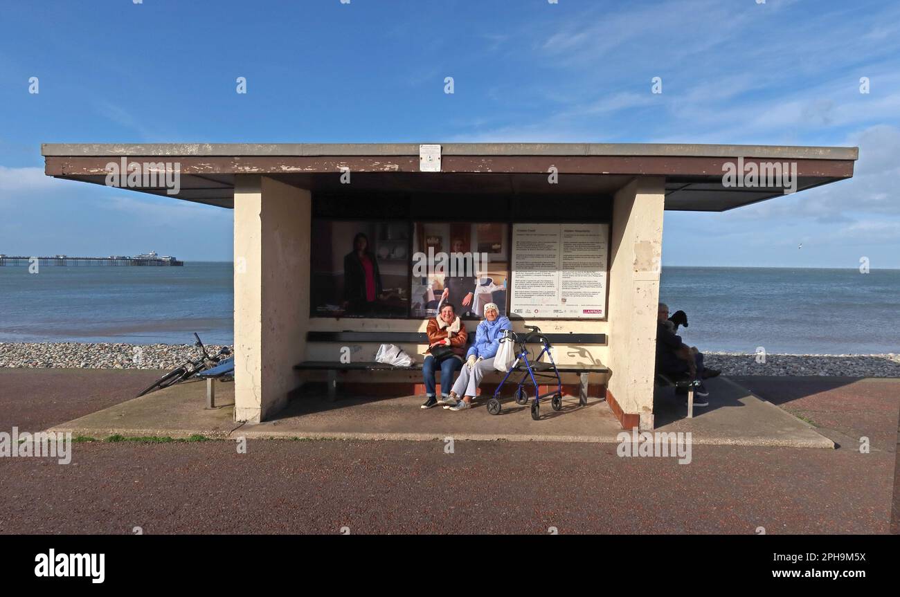 Due Signore che godono di un soleggiato rifugio dal vento, Llandudno Promenade, Conwy, Galles del Nord, Regno Unito, LL30 2LP Foto Stock