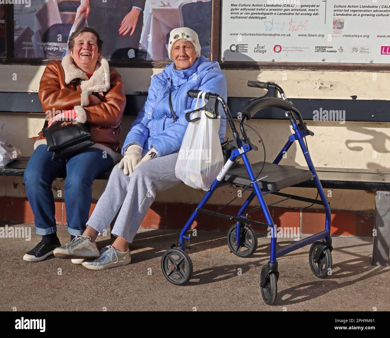 Due Signore che godono di un soleggiato rifugio dal vento, Llandudno Promenade, Conwy, Galles del Nord, Regno Unito, LL30 2LP Foto Stock