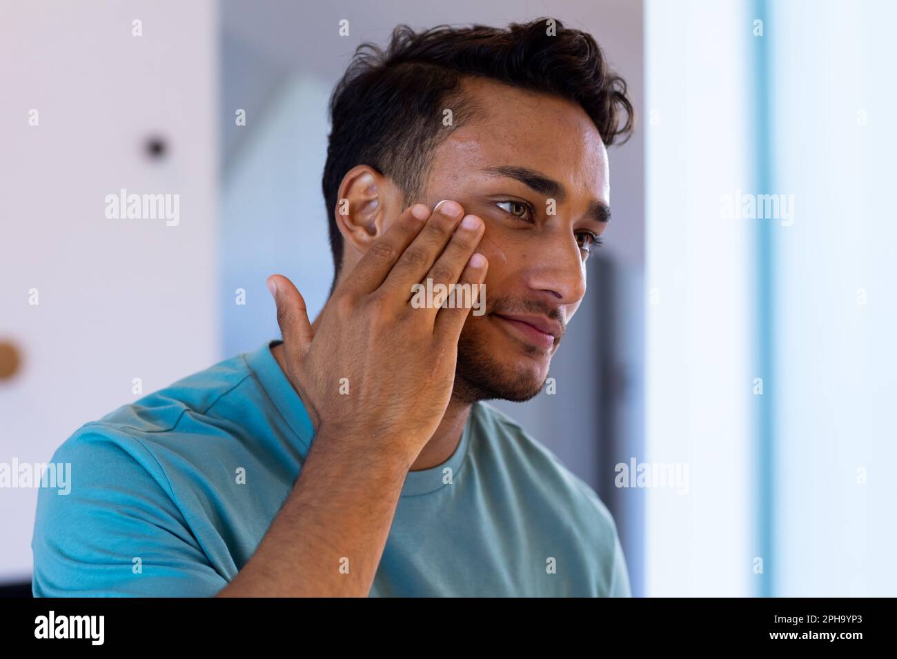 Uomo biraciale sorridente che guarda se stesso nello specchio del bagno e applica crema viso Foto Stock