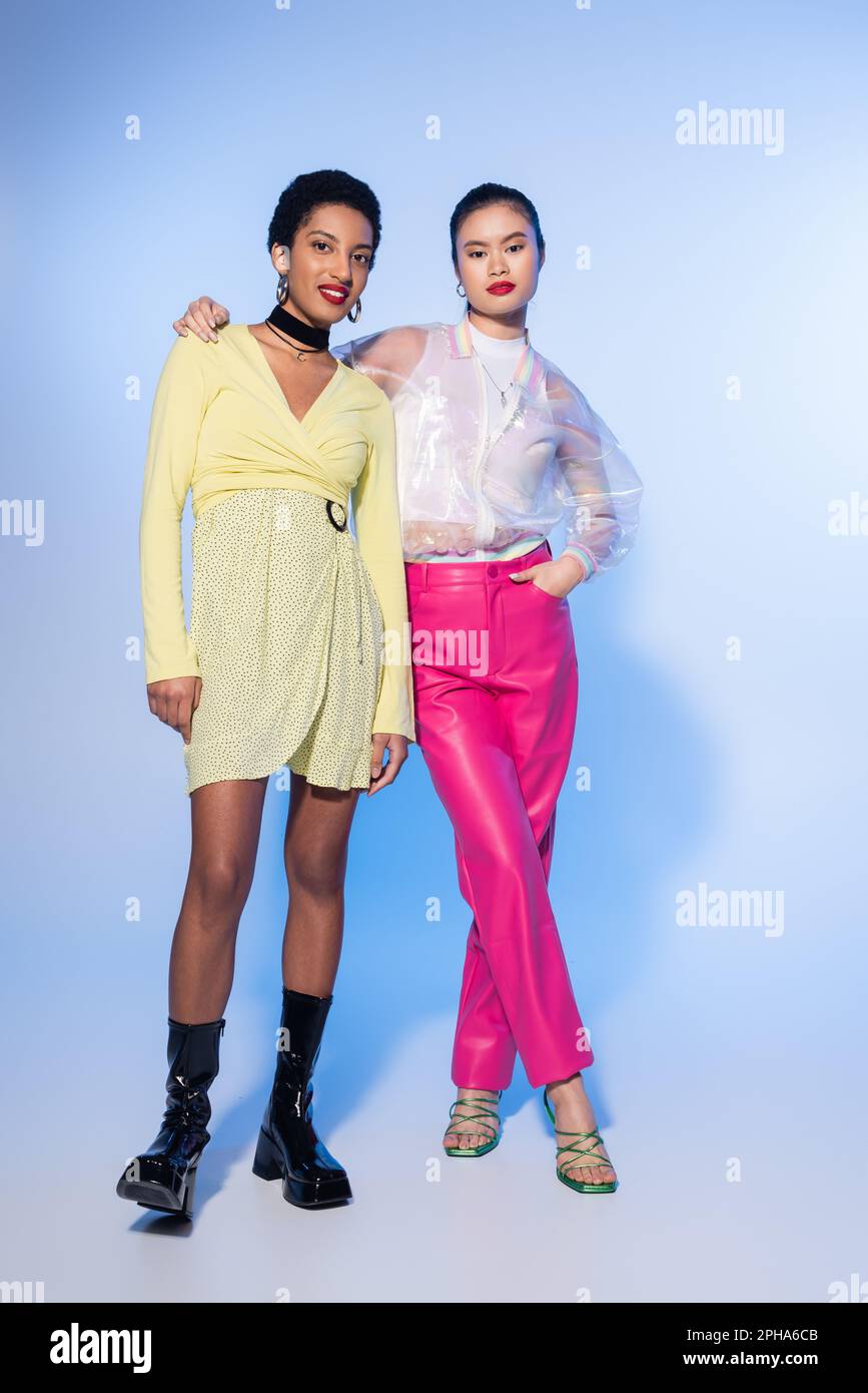 Donna asiatica alla moda che abbraccia sorridente amico afroamericano su sfondo blu, immagine di scorta Foto Stock