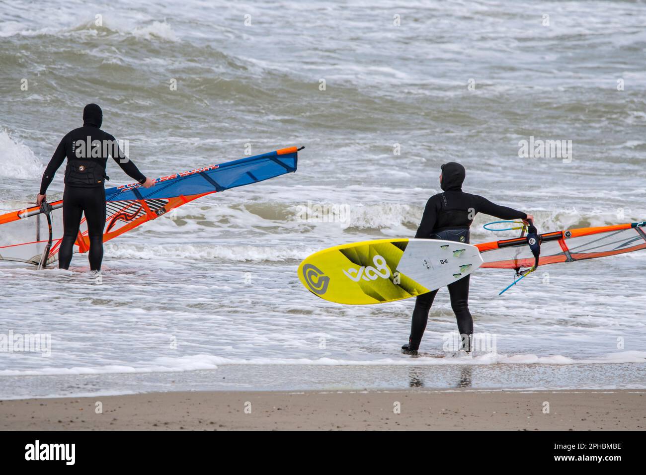 Due windsurf in mute nere che entrano in acqua per praticare il windsurf classico lungo la costa del Mare del Nord in condizioni di vento durante la tempesta invernale Foto Stock