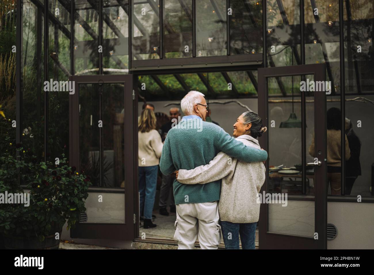 Vista posteriore di amici anziani maschi e femmine in piedi con le braccia intorno all'altro durante la festa a cena Foto Stock