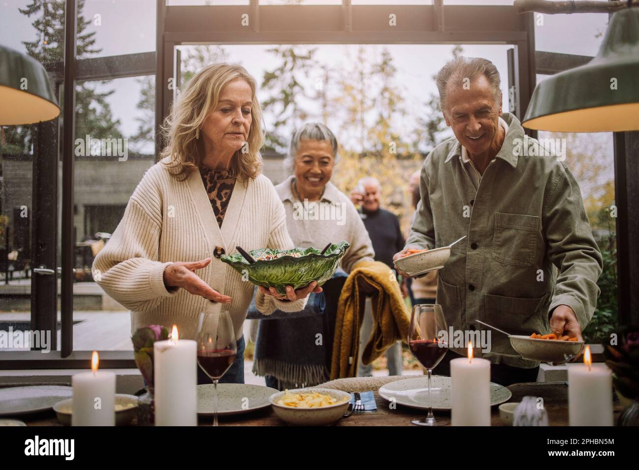 Amici anziani maschili e femminili che si aiutano a vicenda mentre si fissa il tavolo durante la festa della cena Foto Stock