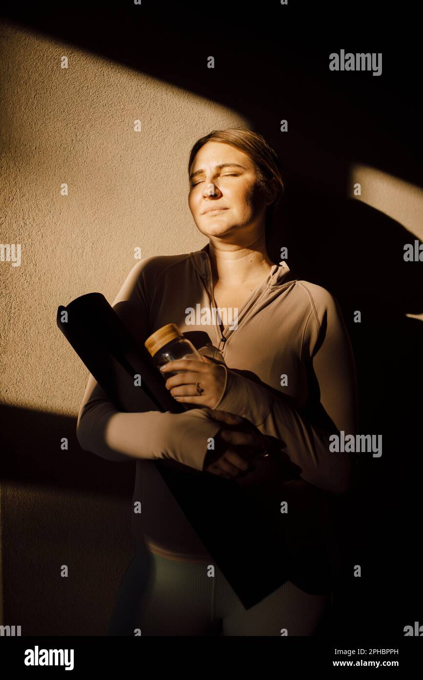 Donna con gli occhi chiusi in piedi contro la parete con luce solare sul viso Foto Stock