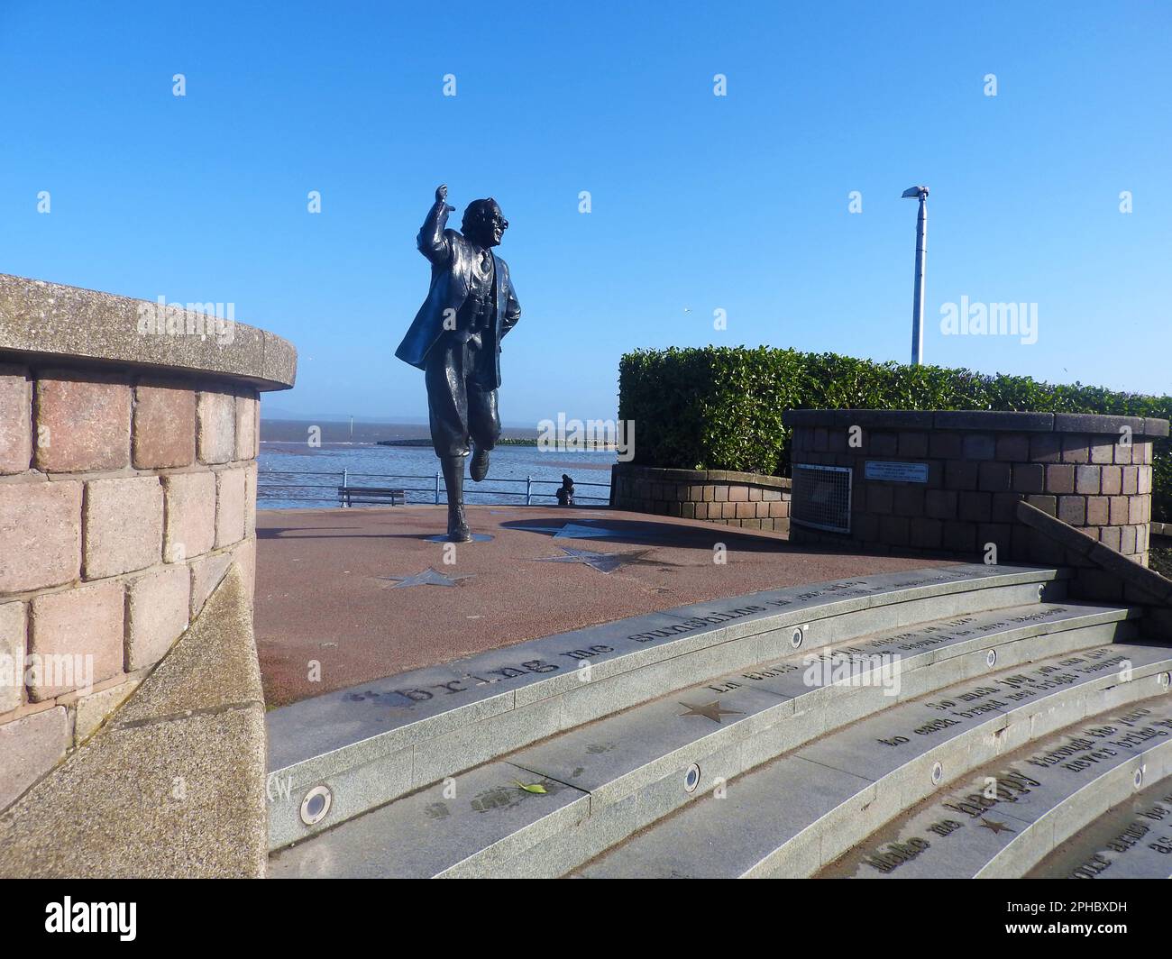 La statua di Eric Morecombe (Eric Bartolomeo. 1926–1984) della prima commedia due Morecambe e Wise. Si trova sul lungomare di Morecambe, Lancashire, Regno Unito, in cima a una serie di gradini incisi con le parole della firma delle coppie "Be me Sunshine" incise in un carattere stile macchina da scrivere. Fu scolpita da Graham Ibbeson e fu svelata dalla defunta regina Elisabetta II nel luglio 1999. Si trova non lontano dalla famosa torre dell'orologio 1905 Torre dell'orologio. Progettato da Cressey e Keighley costruito in mattoni rossi con bande e medicazioni in arenaria. Foto Stock