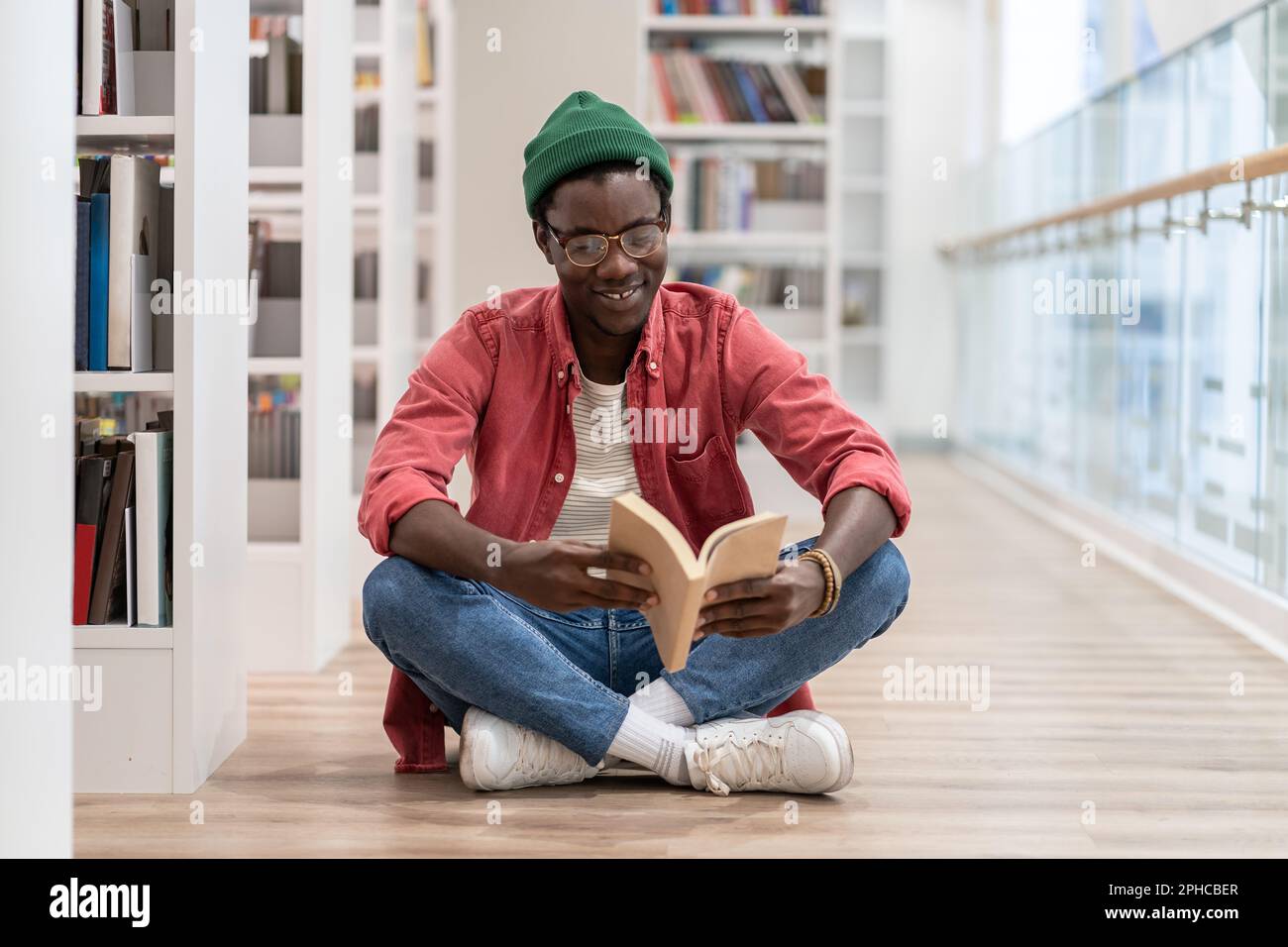 Felice uomo del millennio afroamericano seduto sul pavimento leggendo il libro mentre visita la biblioteca Foto Stock