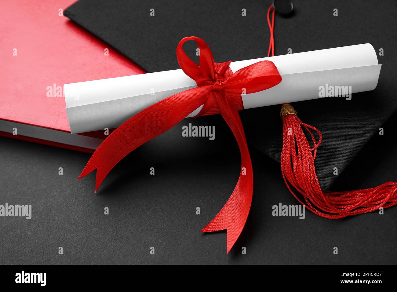 Diploma con nastro rosso, cappello di laurea e libro su sfondo nero Foto Stock