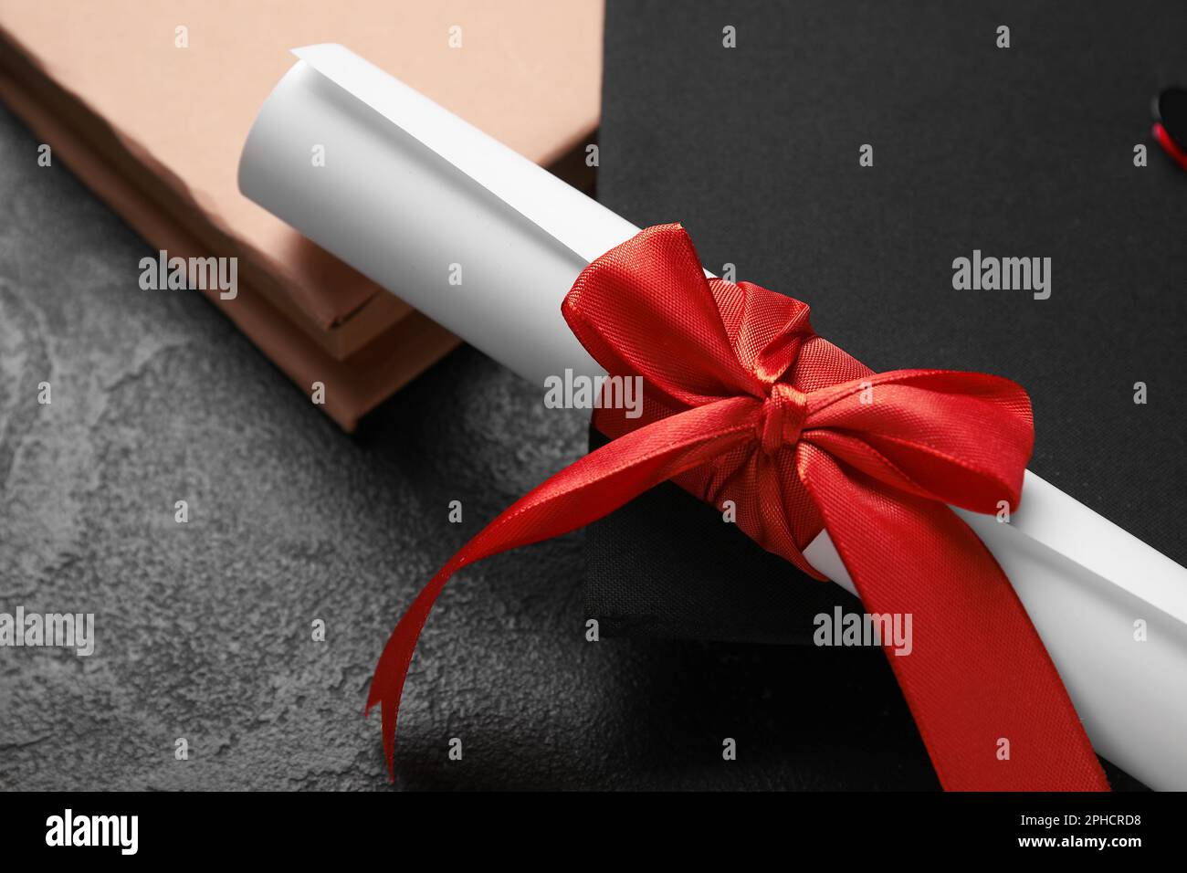 Diploma con nastro rosso, cappello di laurea e libro su tavolo nero Foto Stock