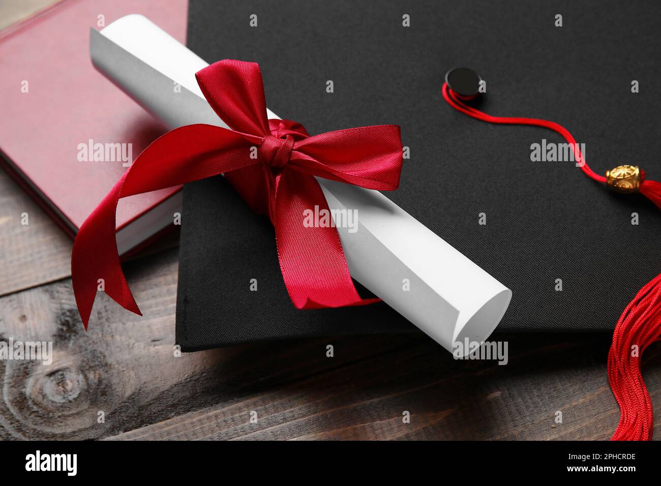 Diploma con nastro rosso, cappello di laurea e libro su tavolo di legno Foto Stock
