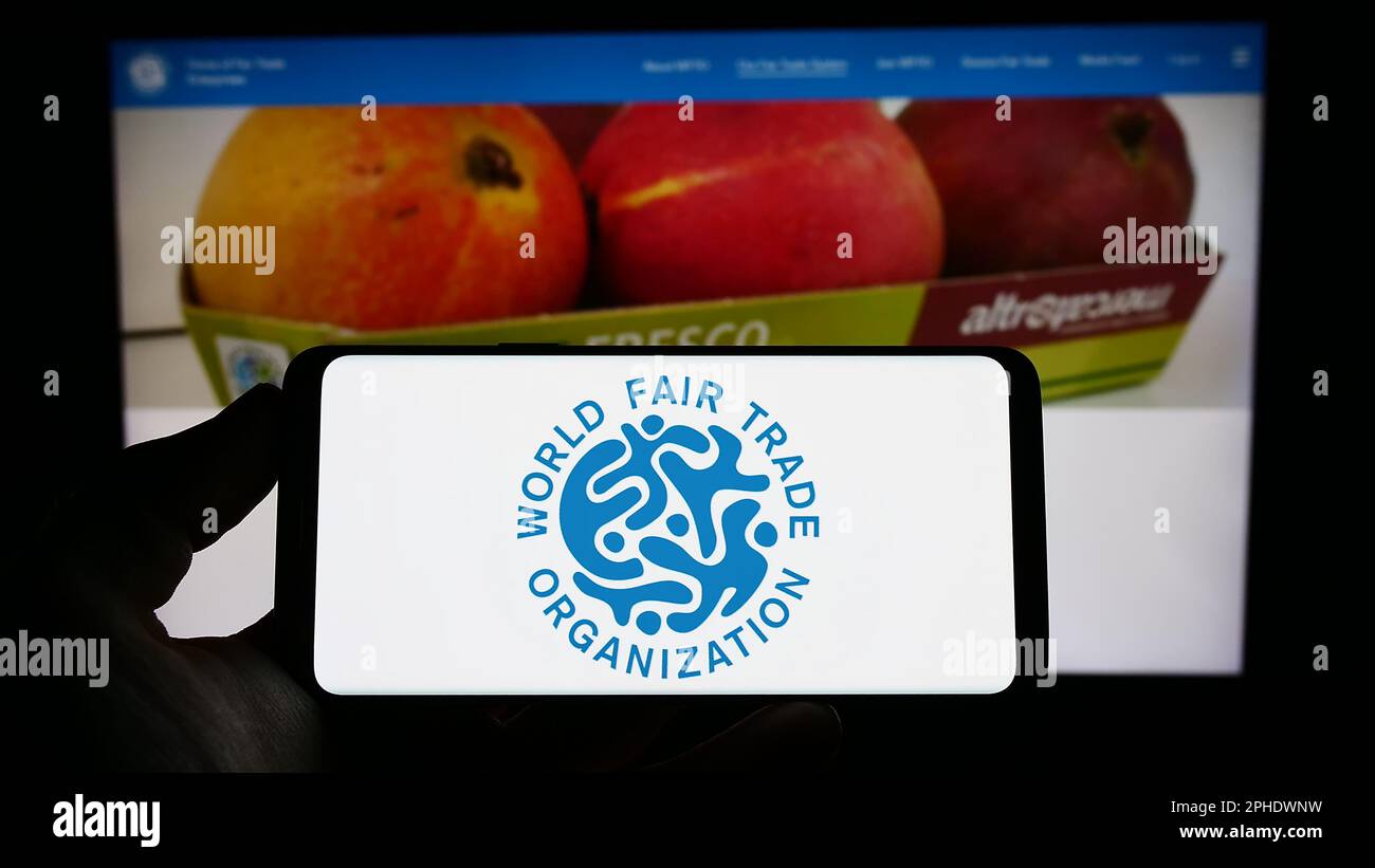 Persona che tiene il cellulare con il logo della World Fair Trade Organization (WFTO) sullo schermo di fronte alla pagina web. Messa a fuoco sul display del telefono. Foto Stock