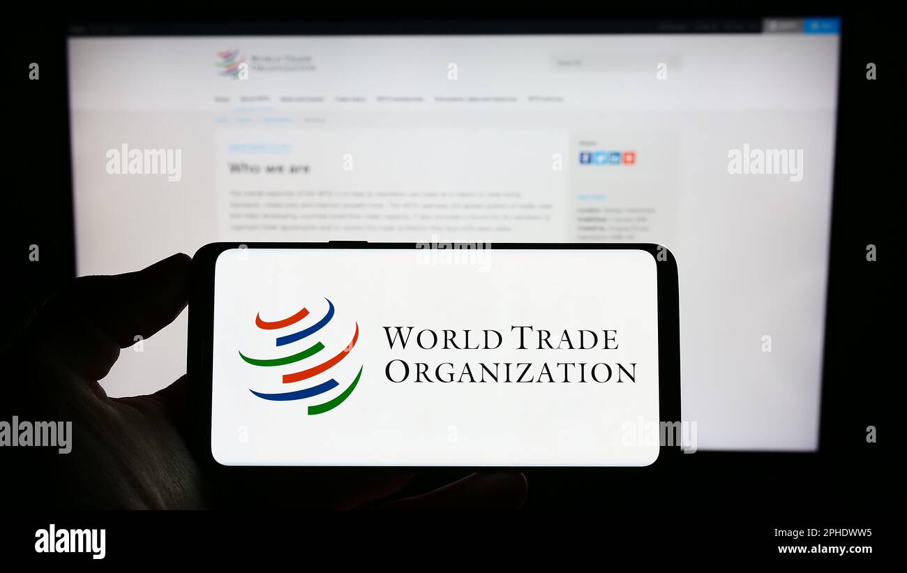 Persona che tiene il cellulare con il logo dell'Organizzazione Mondiale del Commercio (OMC) sullo schermo davanti alla pagina web. Messa a fuoco sul display del telefono. Foto Stock