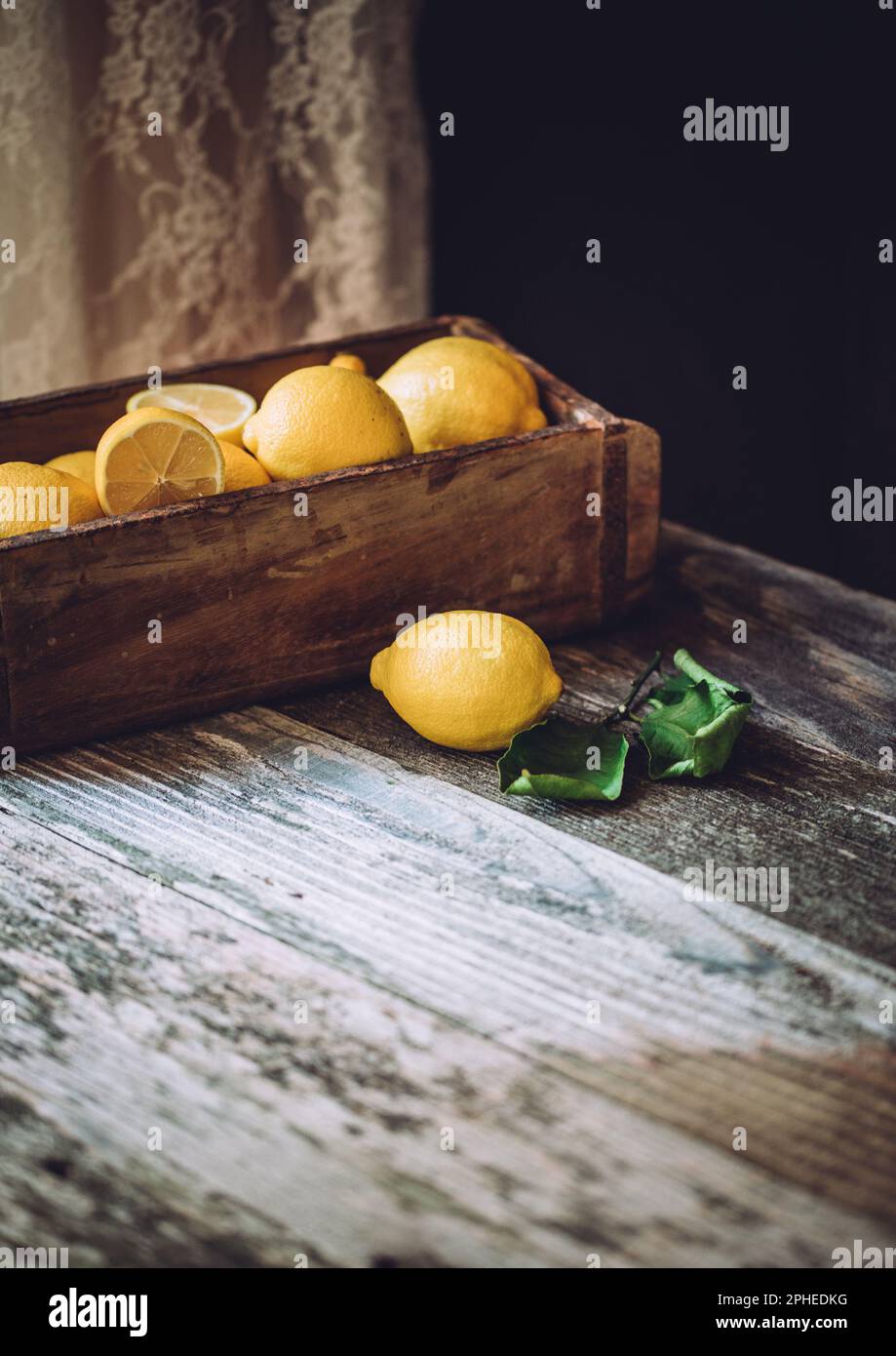 Angolo alto di limoni maturi e pezzi tagliati in scatola di legno messo sul tavolo in luce del giorno contro sfondo scuro in cucina Foto Stock