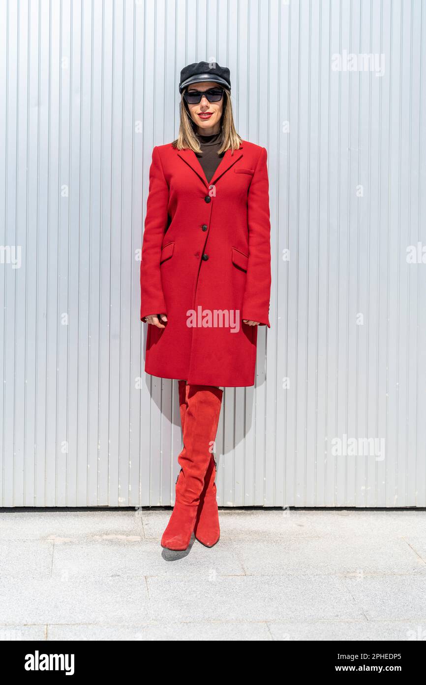 Corpo intero di giovane donna con rossetto rosso in cappotto rosso e cappello sorridente e guardando la macchina fotografica mentre si trova contro la parete grigia Foto Stock