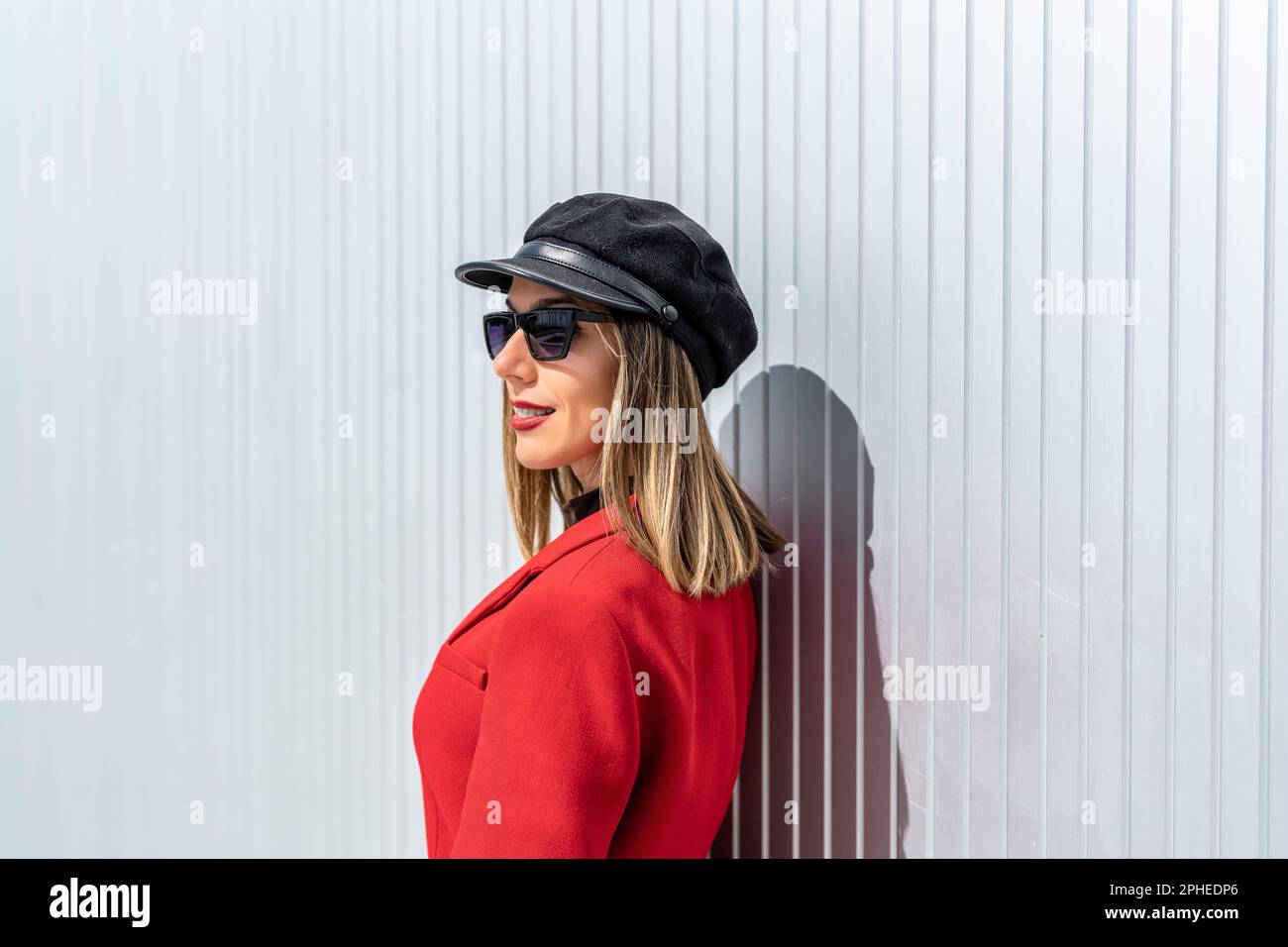 Vista laterale della giovane donna con rossetto rosso in cappotto rosso e cappello sorridente e guardando la macchina fotografica mentre si trova contro la parete grigia Foto Stock