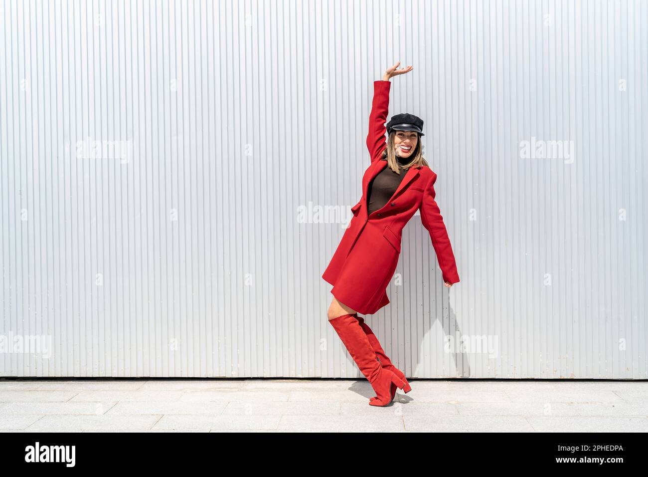 Corpo pieno di giovane donna con cappello e rossetto rosso sorridente e posante in piedi sui tiptoes vicino alla parete grigia Foto Stock