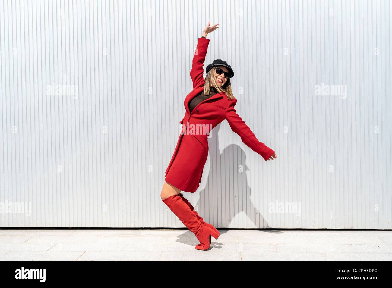 Corpo pieno di giovane donna con cappello e rossetto rosso sorridente e posante in piedi sui tiptoes vicino alla parete grigia Foto Stock