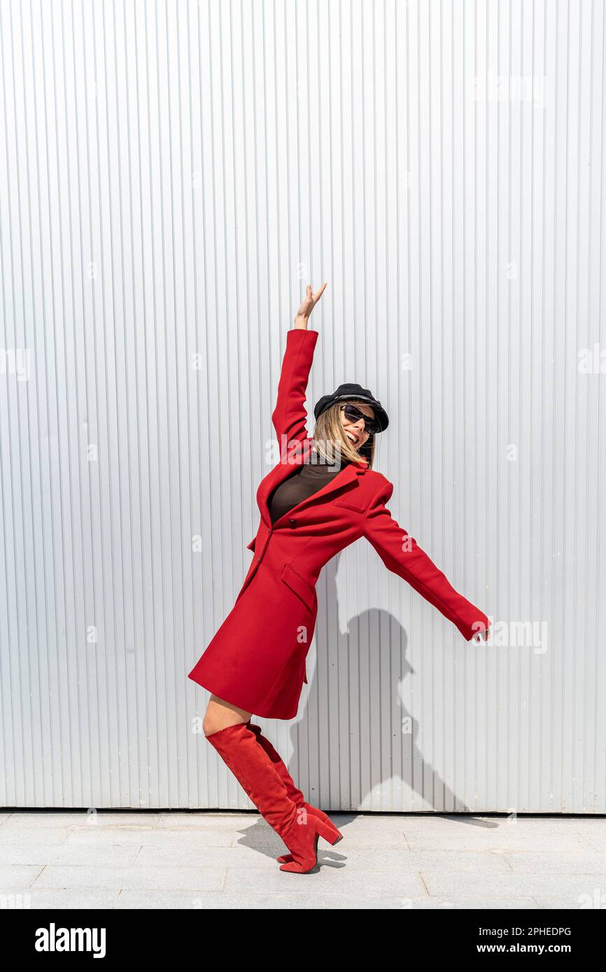 Corpo pieno di giovane donna con cappello e rossetto rosso sorridente e posante in piedi sui tiptoes vicino alla parete grigia Foto Stock