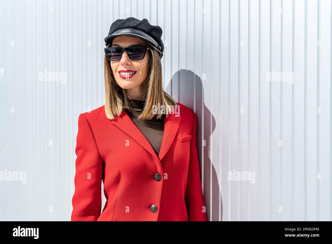 Giovane donna con rossetto rosso in cappotto rosso e cappello sorridente e guardando la macchina fotografica mentre si trova contro la parete grigia Foto Stock