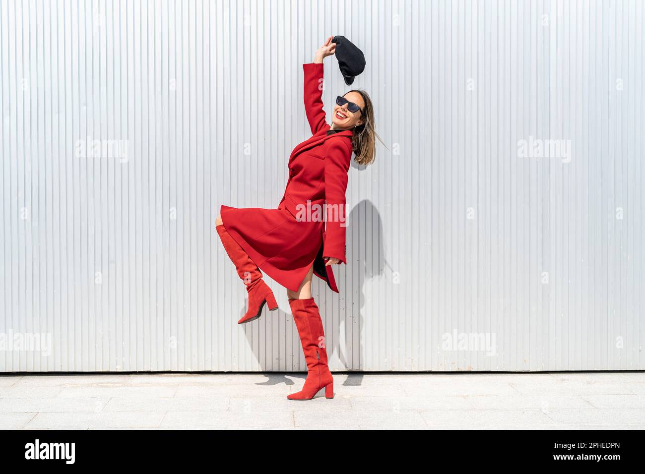Corpo pieno di giovane donna sorridente e in posa con una gamba in su mentre tiene il cappello sopra la testa vicino alla parete grigia Foto Stock