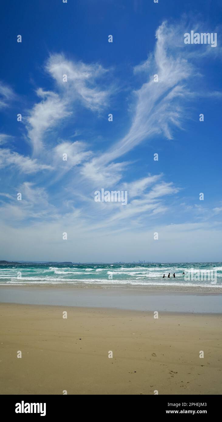Surf spiaggia in una giornata ventosa. Vista dalla spiaggia sabbiosa sopra il surf allo skyline della città all'orizzonte, con le nuvole di cirrus spazzate dal vento contro un cielo blu. Foto Stock