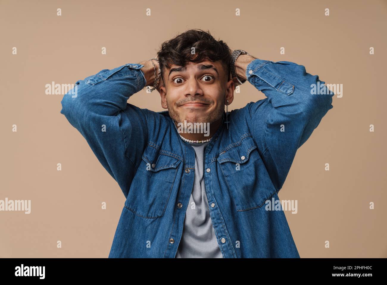 Giovane uomo elegante con labbra a portafoglio che tiene la testa e guarda la macchina fotografica , mentre si trova su un isolato sfondo marrone Foto Stock