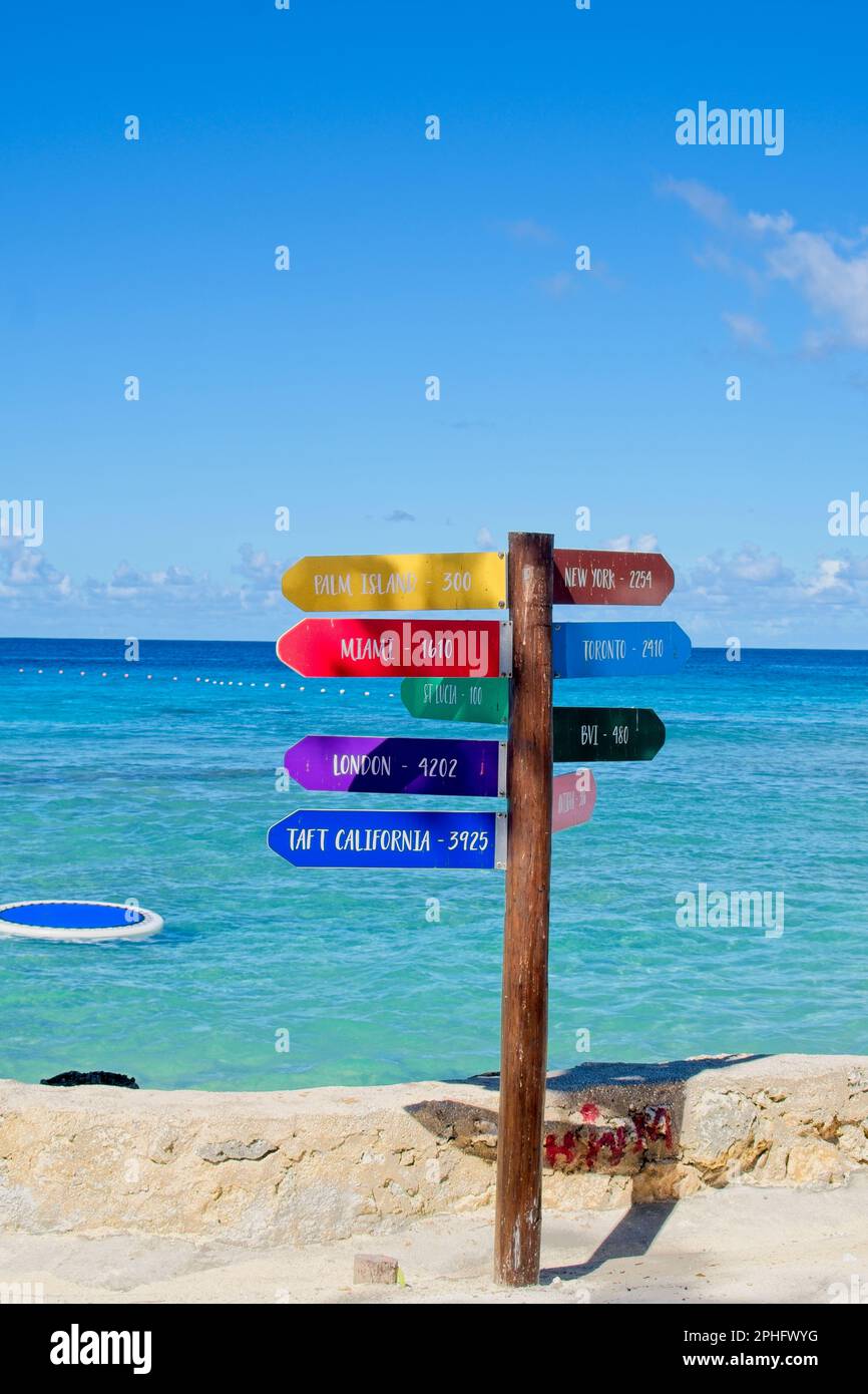15Feb2023 Barbados Caraibi cartellonistica multicolore sul fronte spiaggia che indica le distanze da terre lontane. Cielo azzurro e mare calmo. Foto Stock