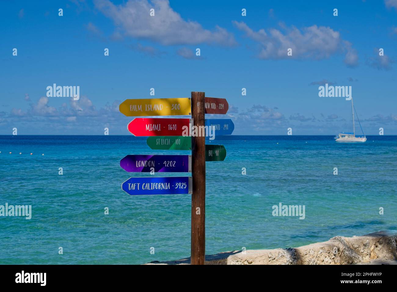15Feb2023 Barbados Caraibi cartellonistica multicolore sul fronte spiaggia che indica le distanze da terre lontane. Cielo azzurro e mare calmo. Foto Stock