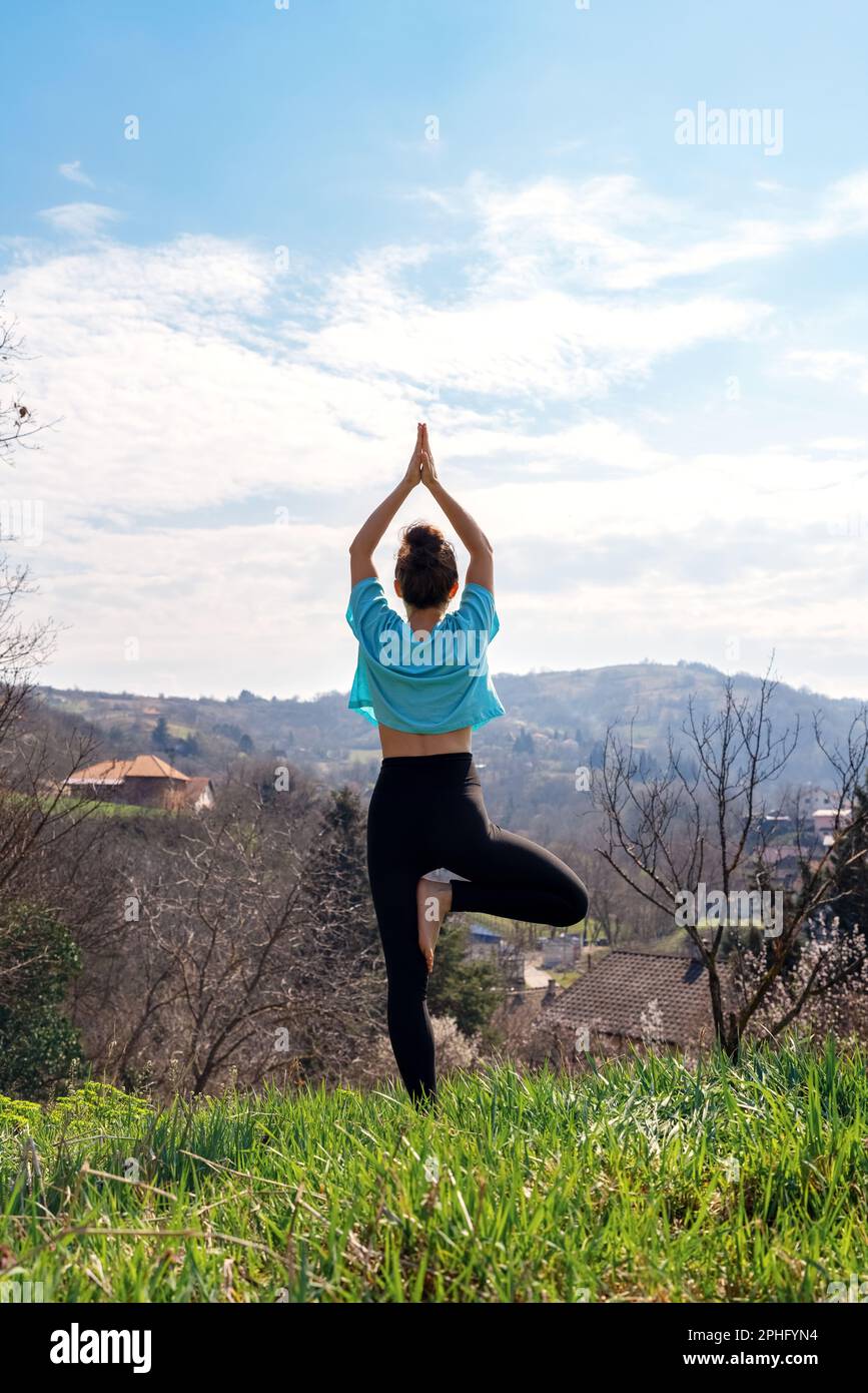 Vista posteriore donna che fa yoga esercizio Vrikshasana albero posa in natura contro la montagna. Foto Stock