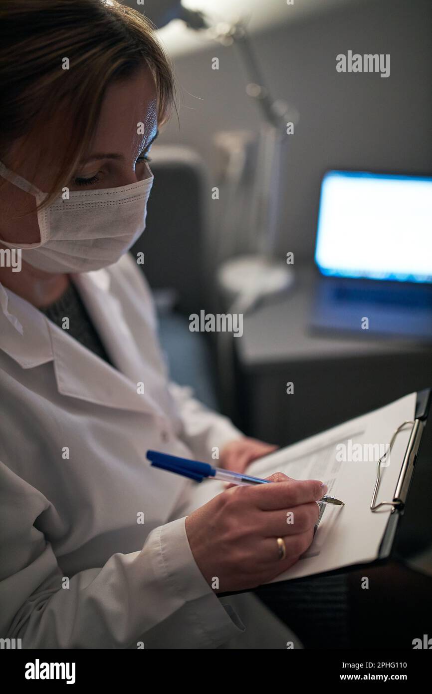Medico che compila un documento. Personale ospedaliero che lavora in servizio notturno. Donna che indossa uniforme, cappuccio e maschera facciale per prevenire l'infezione da virus Foto Stock