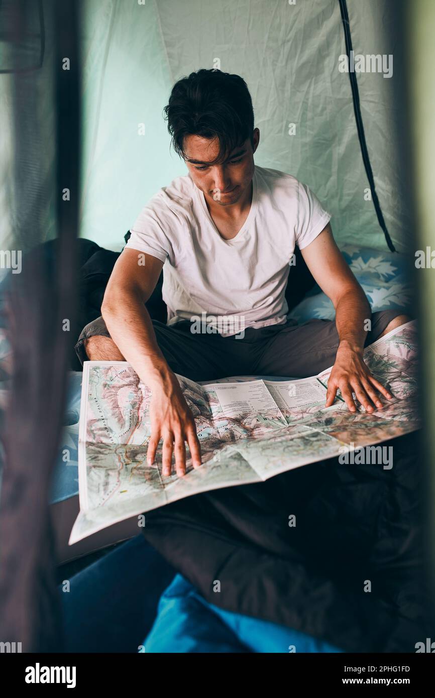 Giovane uomo che guarda una mappa dei sentieri di montagna che pianifica il prossimo viaggio in vacanza estiva seduto in una tenda in campeggio. Concetto di vita del campo Foto Stock