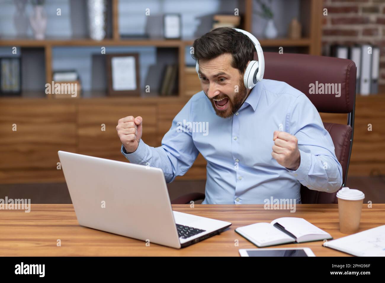 Uomo d'affari maturo in camicia e cuffie che guarda la partita sportiva sul posto di lavoro all'interno dell'ufficio, l'uomo che fa il tifo per divertirsi sul lavoro, guardando video sul laptop. Foto Stock