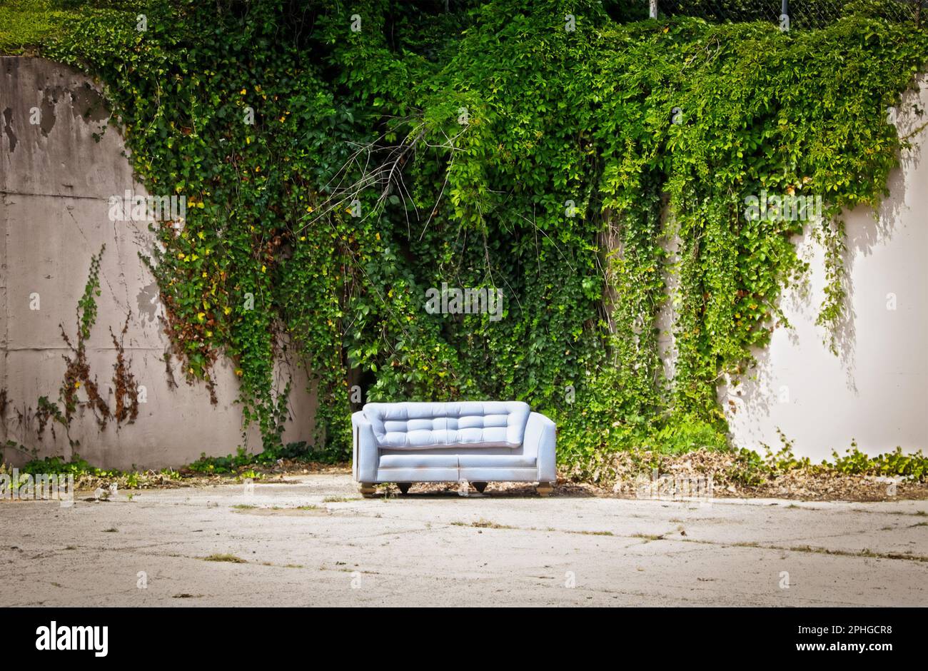 Vecchio divano abbandonato seduto in un angolo di parcheggio in cemento con alto muro di cemento coperto di viti sullo sfondo - sembra un set di photoshoot Foto Stock