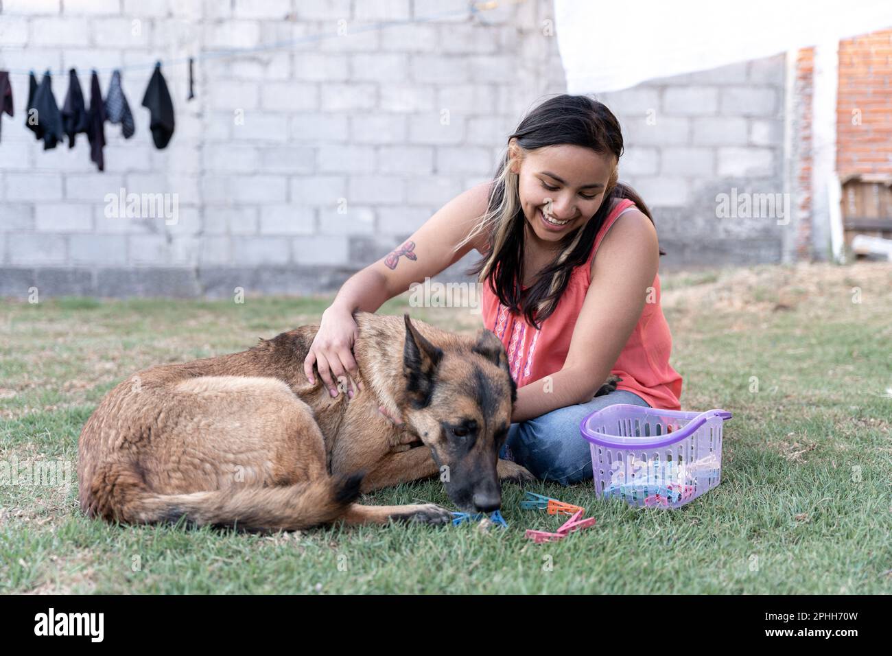 Una giovane donna ispanica sta giocando con il suo cane e i vestiti nel giardino mentre fa il bucato Foto Stock