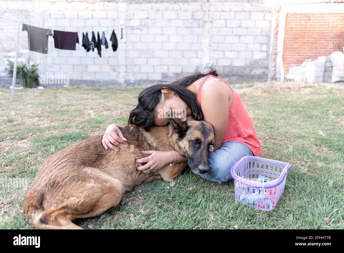 Una giovane donna triste ispanica sta abbracciando il suo cane che viene consolato nel giardino mentre fa il bucato Foto Stock