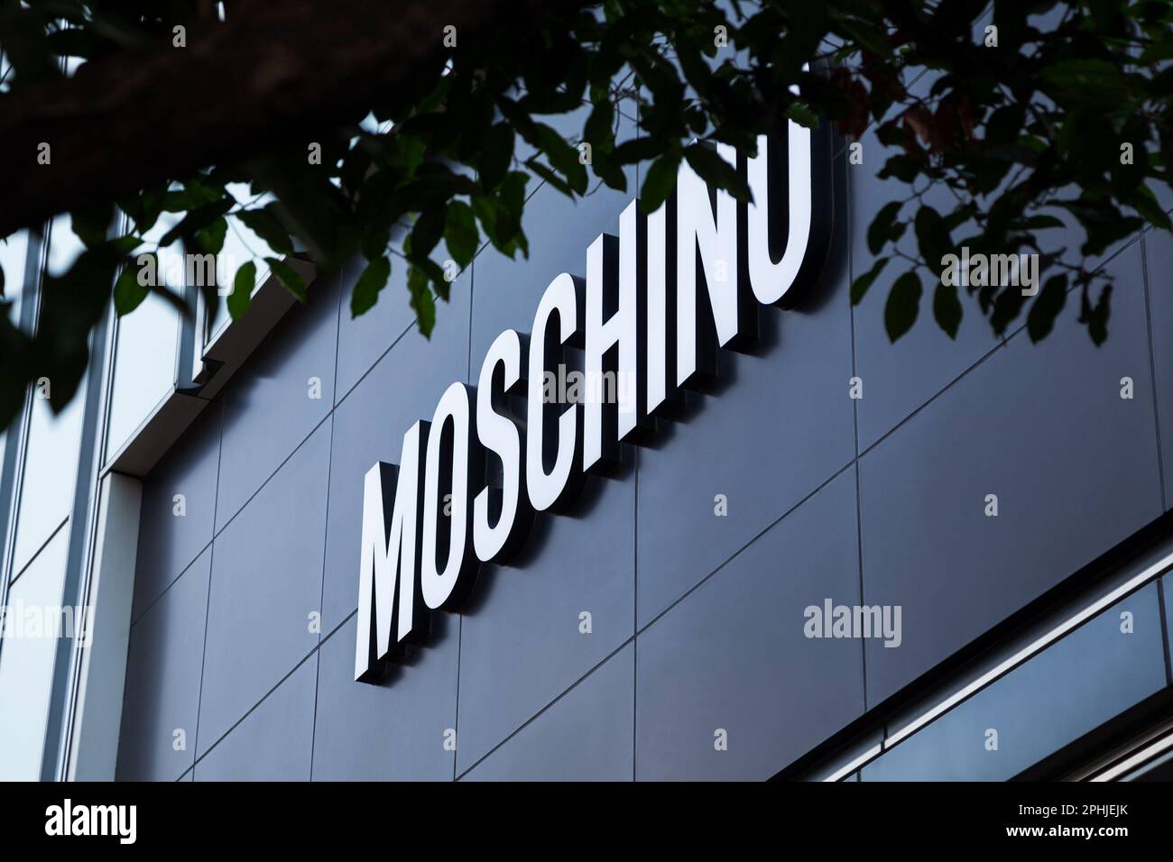 Ho Chi Minh City, Vietnam - 28 marzo 2023: Logo Moschino su una facciata del negozio di moda visto attraverso il fogliame dell'albero. Uscita del marchio di accessori di lusso più diffuso Foto Stock