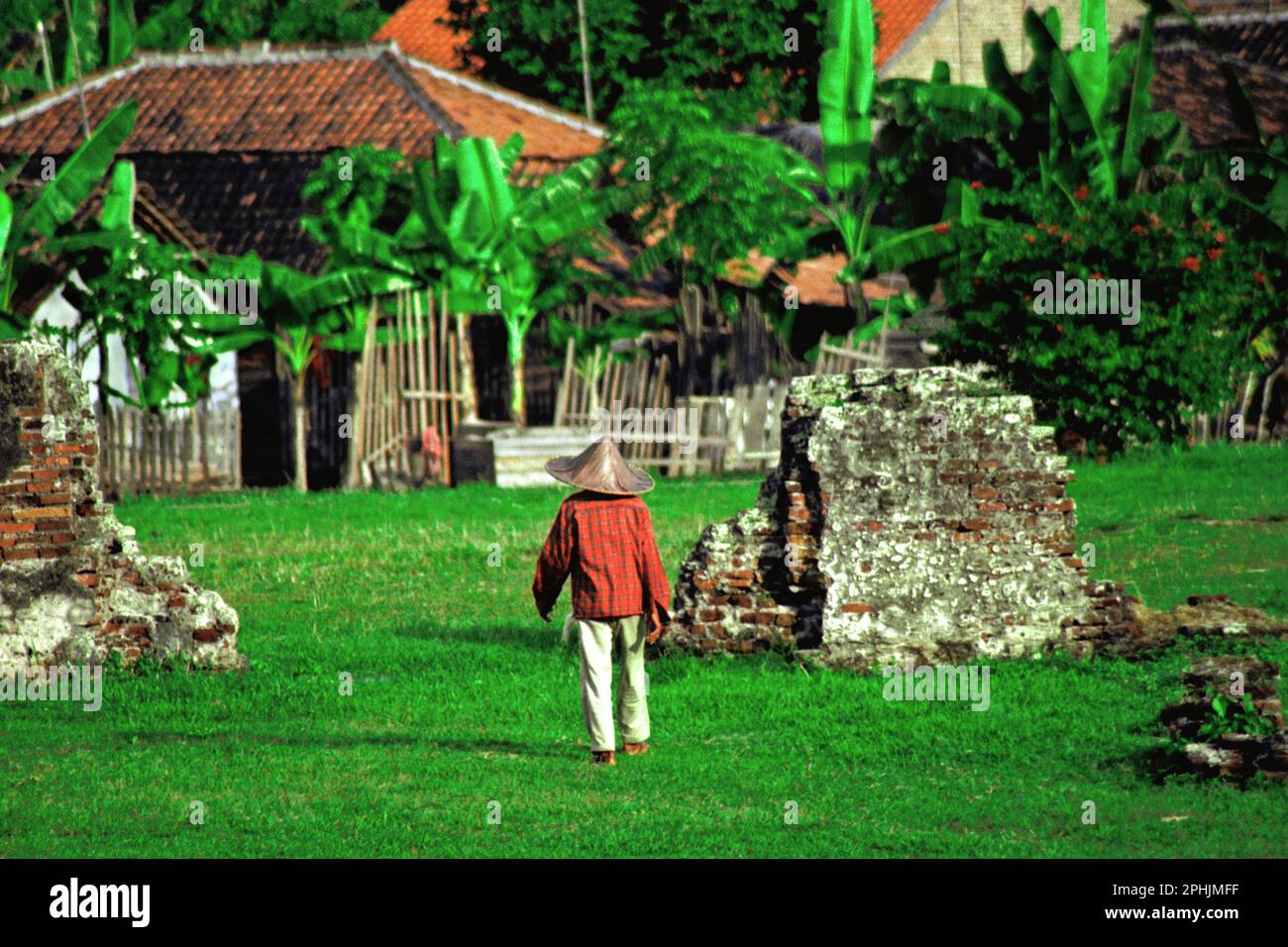 Un villager che indossa il cappello di contadino sta camminando sul campo d'erba vicino alla rovina di un edificio al palazzo di Kaibon, uno degli oggetti del patrimonio culturale del periodo di Banten Sultanato situato in un'area ora chiamata Banten lama (Old Banten) a Serang, Banten, Indonesia. "La sostenibilità del patrimonio culturale è fortemente legata all'effettiva partecipazione delle comunità locali alla conservazione e alla gestione di queste risorse", secondo un team di scienziati guidati da Sunday Oladipo Oladeji nel loro articolo di ricerca pubblicato su Sage Journals il 28 ottobre 2022. Foto Stock
