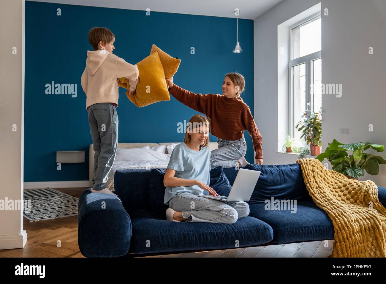 Allegri fratelli europei che combattono il cuscino di un ragazzo e di una ragazza distrarre la mamma affollata che si rilassa sul divano di casa Foto Stock