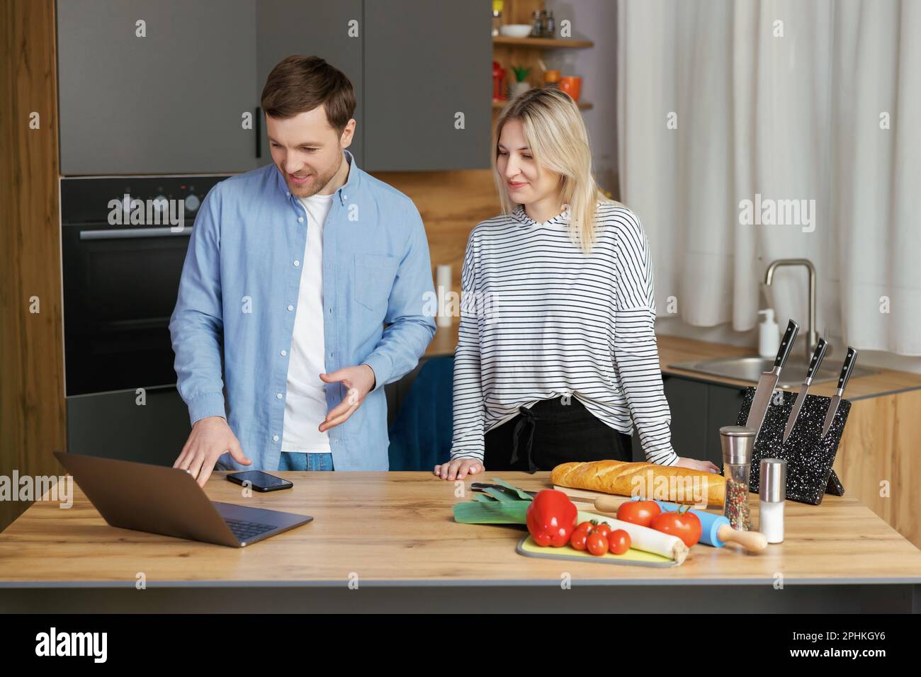 Piacevole coppia di famiglia in piedi vicino a un grande tavolo di legno in una cucina moderna, guardando lo schermo del computer portatile. Felice giovane coppia che inizia a preparare il pranzo Foto Stock